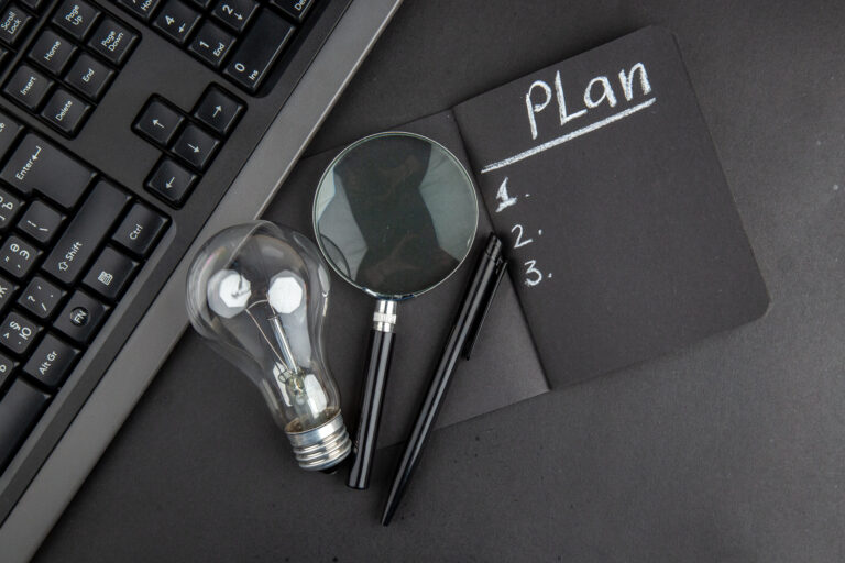 Business Continuity Plan – Notizbuch mit Plan, Glühbirne, Lupe und Tastatur auf schwarzem Schreibtisch