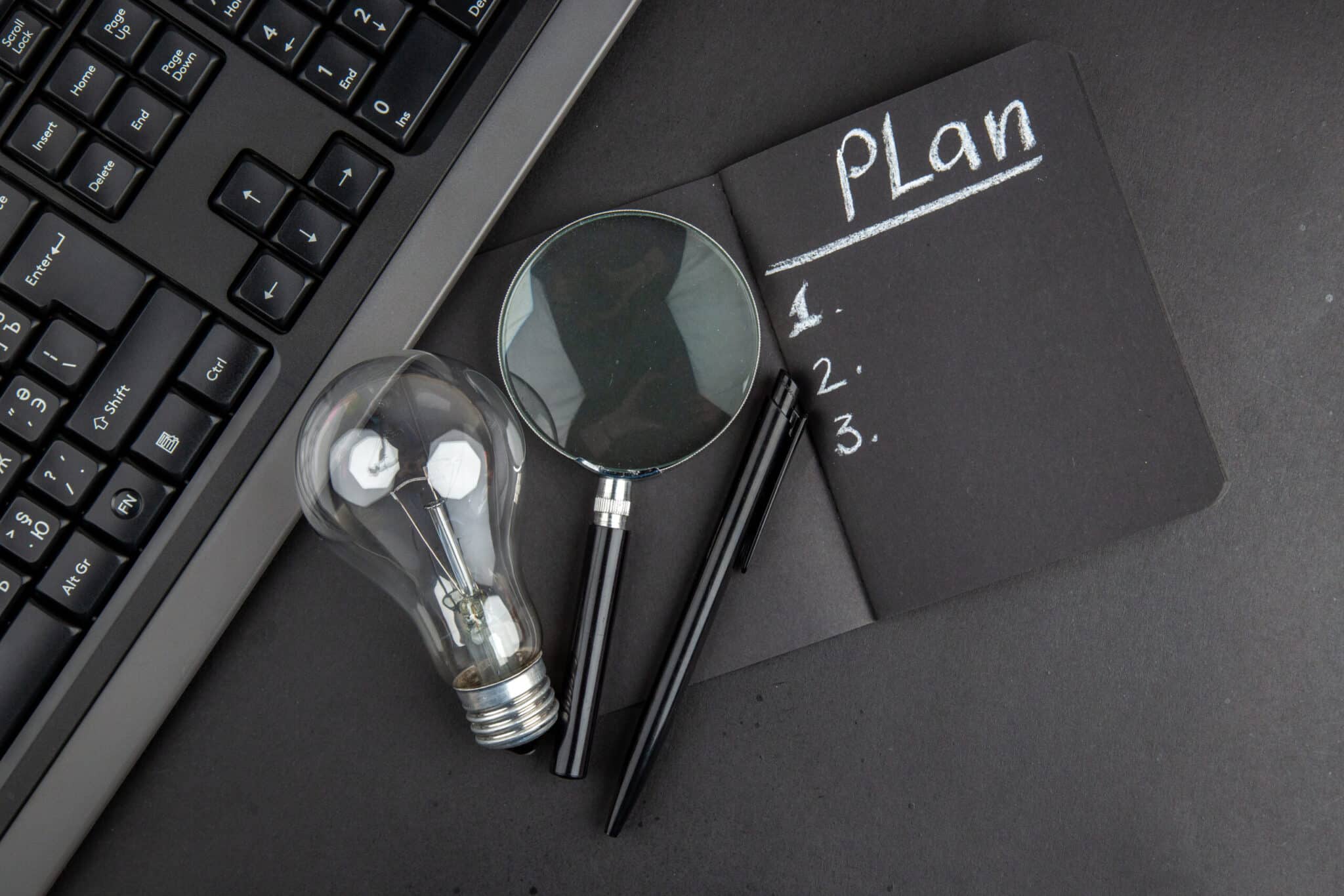 Business Continuity Plan – Notizbuch mit Plan, Glühbirne, Lupe und Tastatur auf schwarzem Schreibtisch