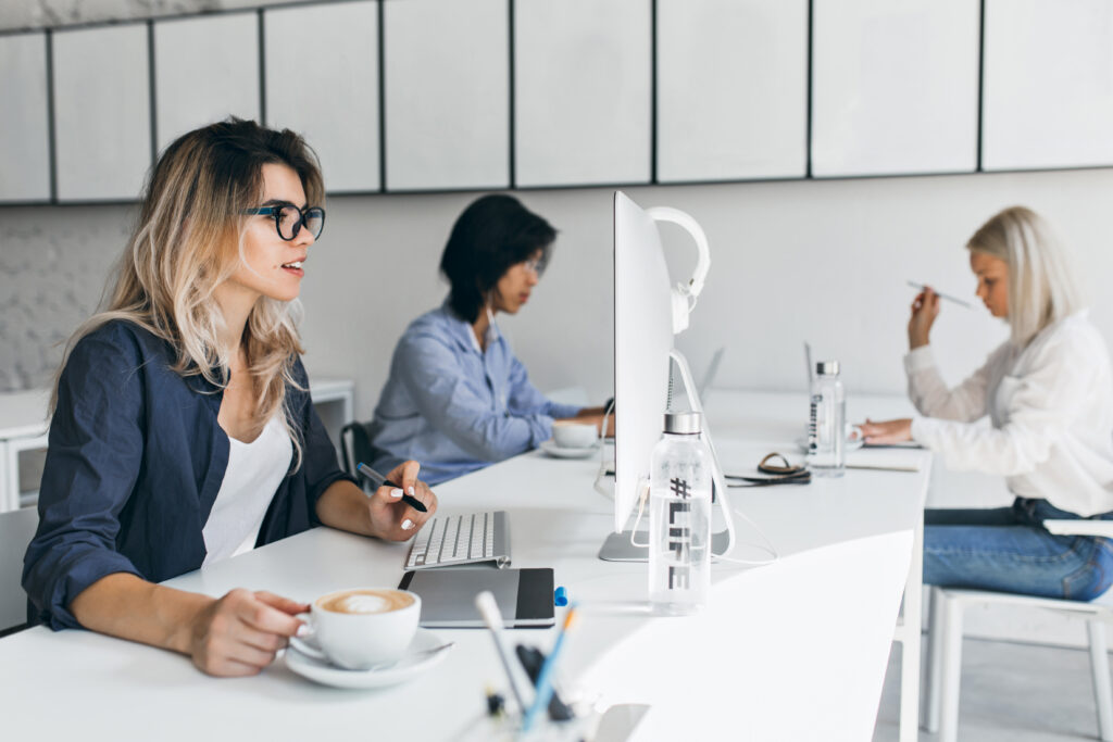 Junge Frau mit Kaffee arbeitet konzentriert an einem Bildschirm im offenen Workspace – Beispiel für moderne Arbeitskultur