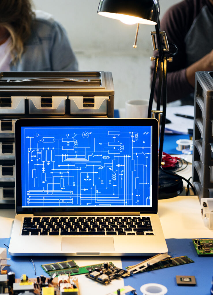 Laptop mit technischem Schaltplan-Blueprint auf Bildschirm, umgeben von Hardware-Teilen